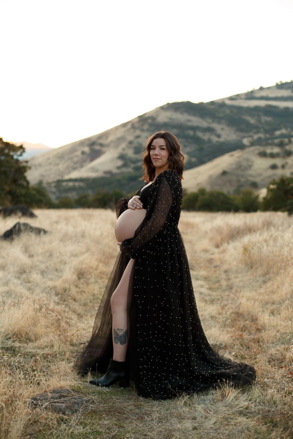 A pregnant woman poses in a flowing black sheer gown outdoors in a golden field during sunset with rolling hills behind her.