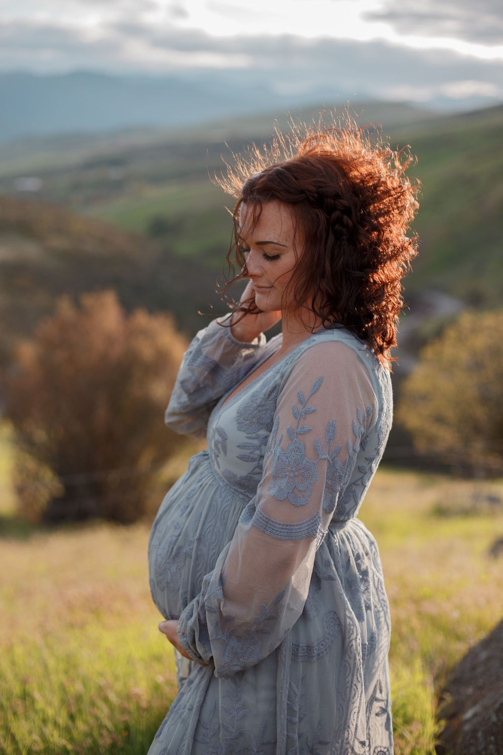 A pregnant woman in a flowing dress stands in golden sunlight on a hillside with mountains in the background.