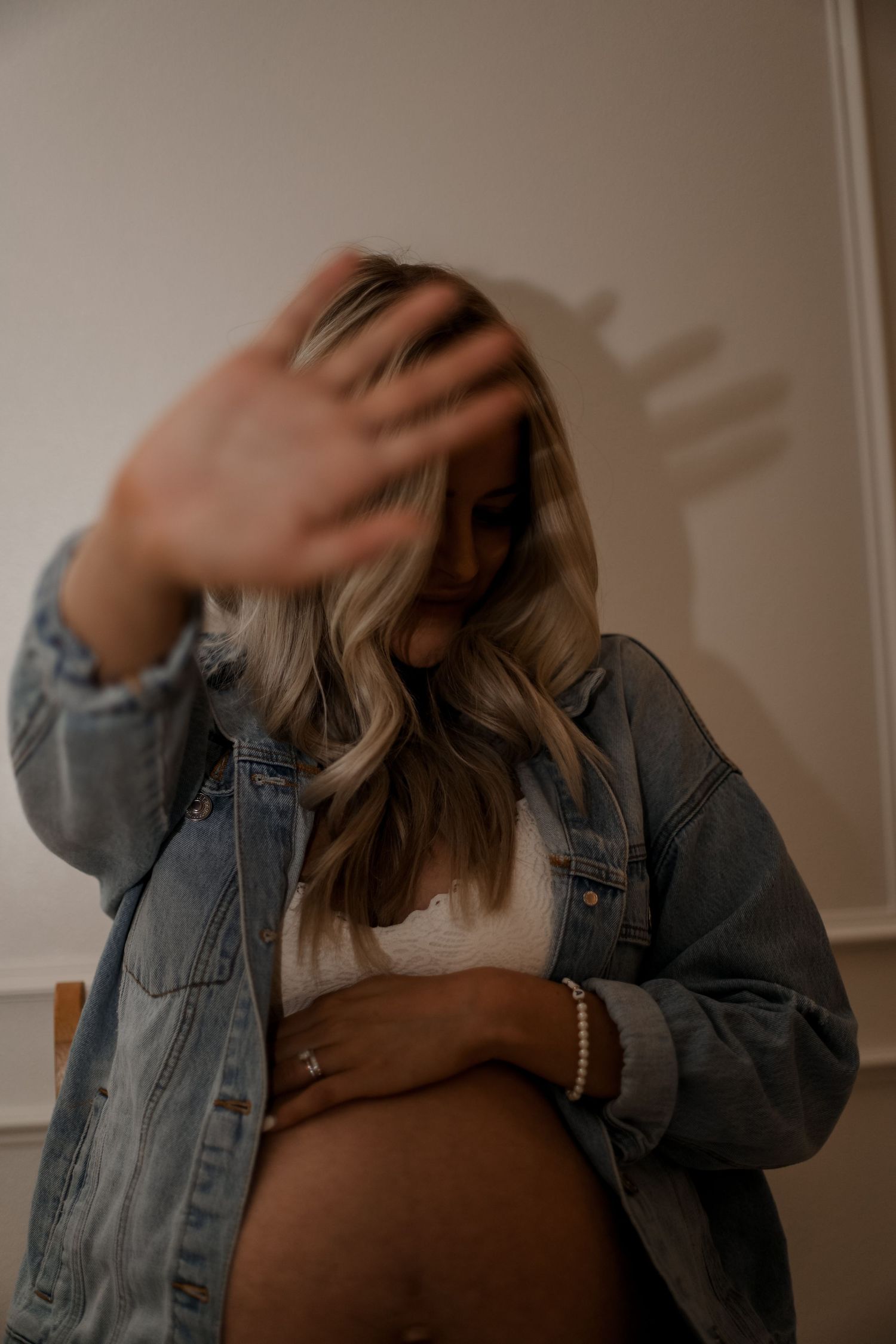 A maternity photo showing someone in a denim jacket with wavy hair casting shadows on a white wall.