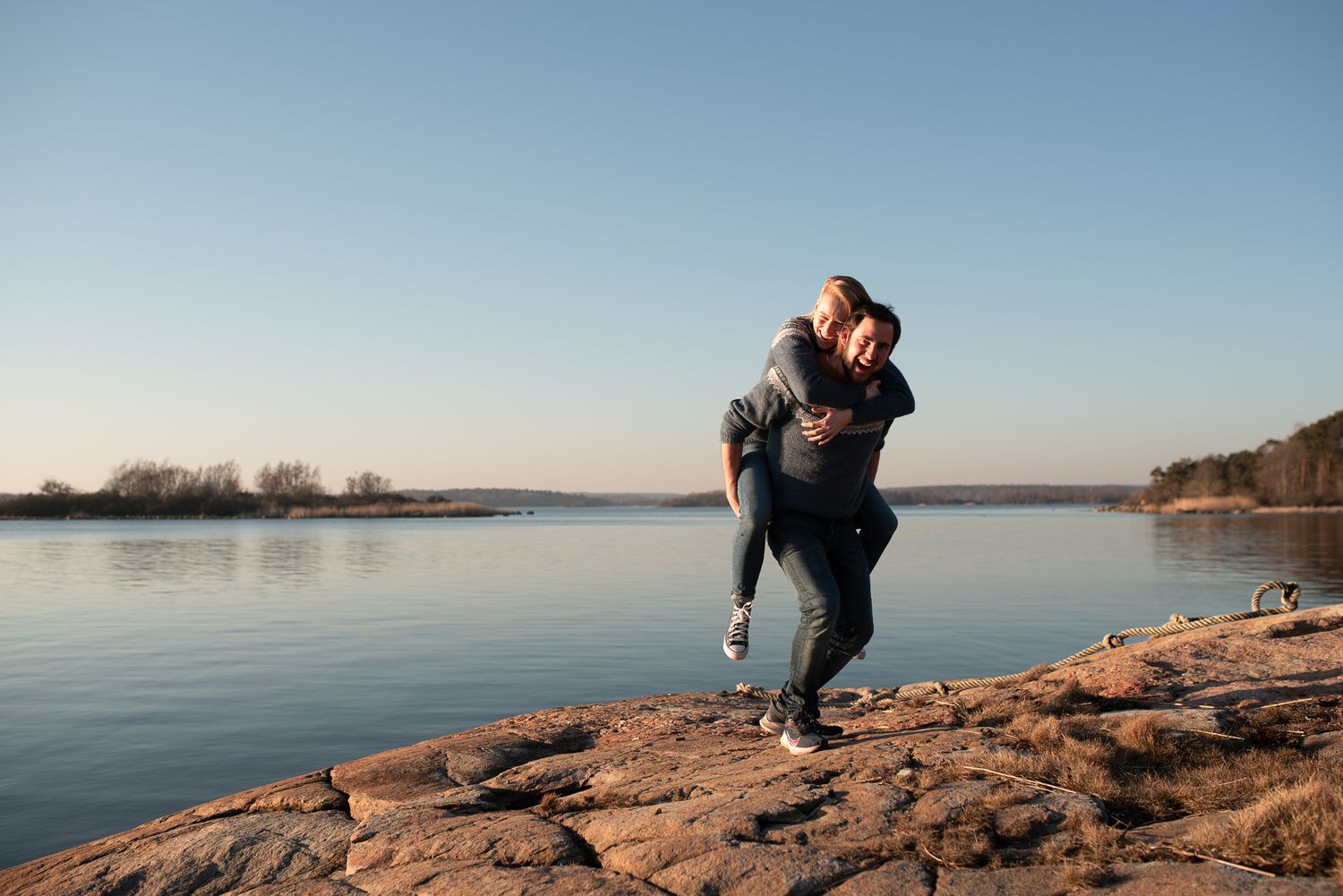 Couple embracing on rocky shoreline against calm water and sunset sky.