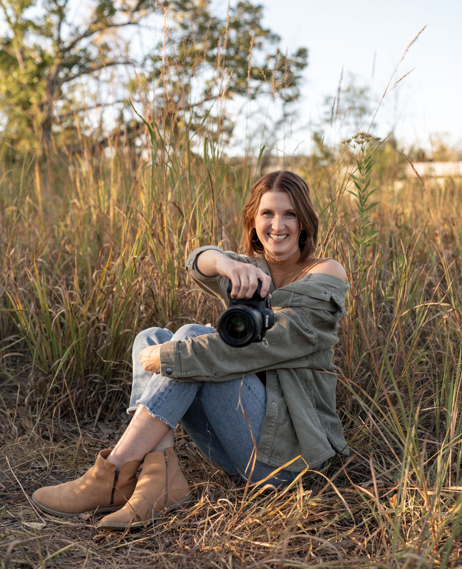 Photographer with camera sits in tall autumn grass wearing casual attire and brown boots.