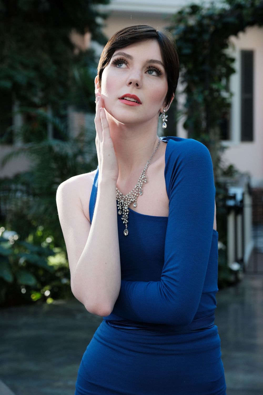 Brunette woman in royal blue dress with silver necklace looking up outdoors.
