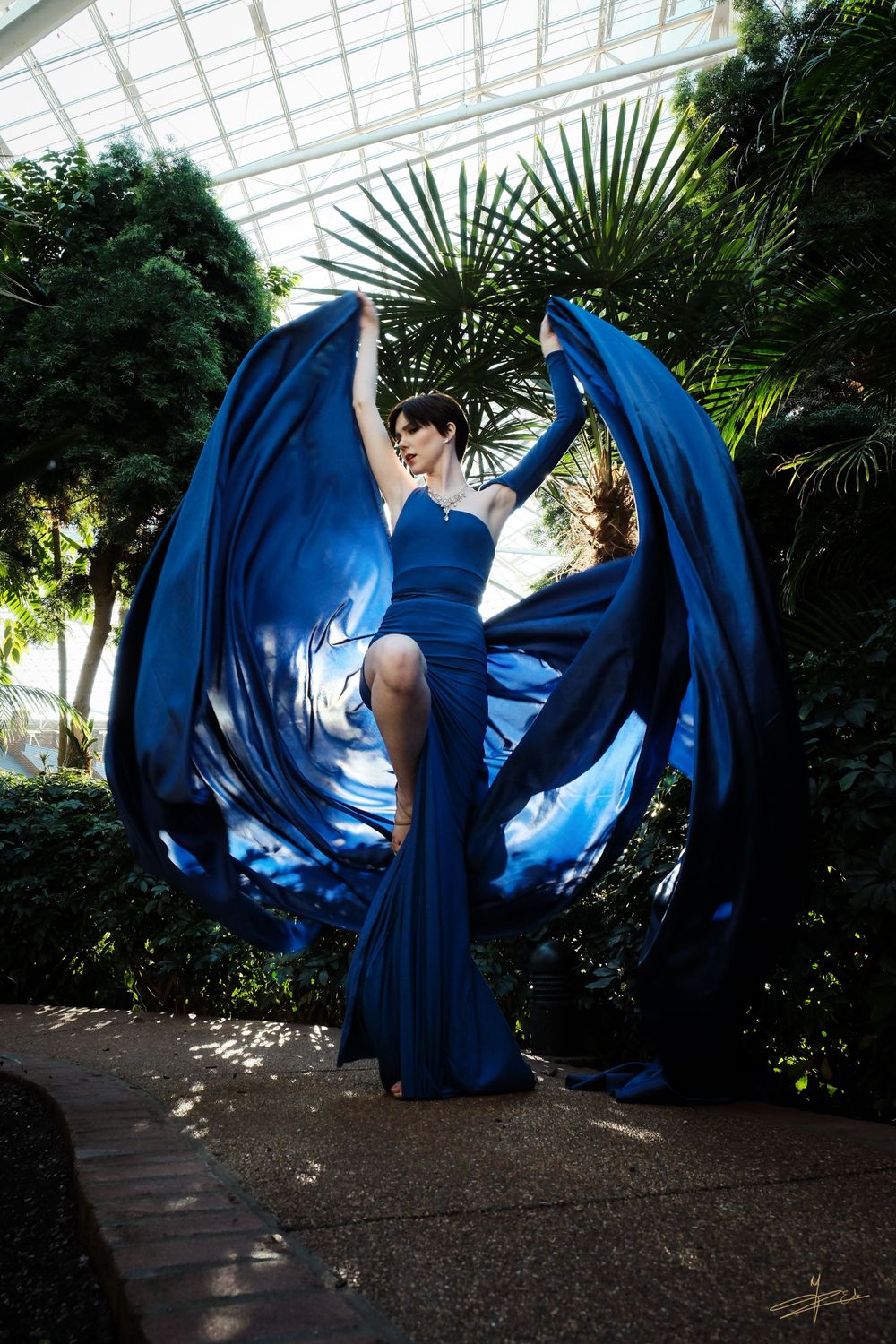 Woman in flowing blue gown with dramatic fabric wings spread wide in tropical greenhouse.