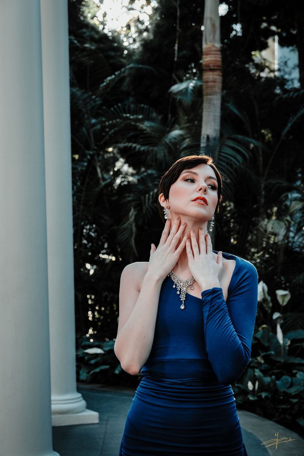 Woman in blue one-shoulder dress with diamond jewelry posing among tropical plants.
