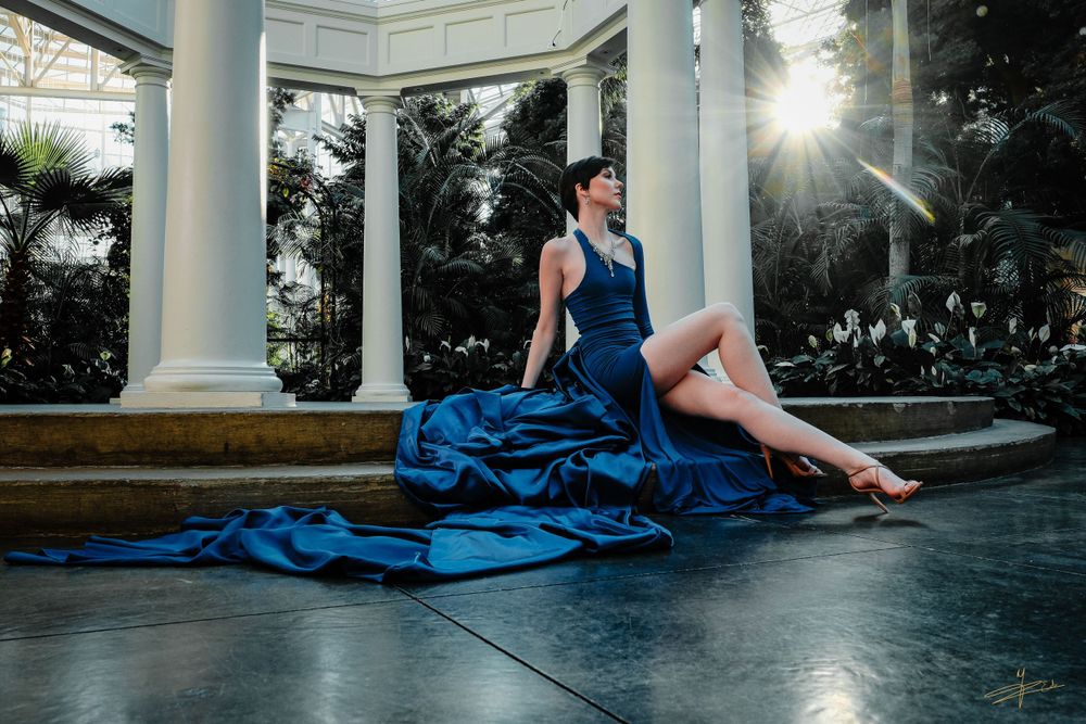 Elegant woman in blue gown seated on floor beside white columns in sunlit conservatory.
