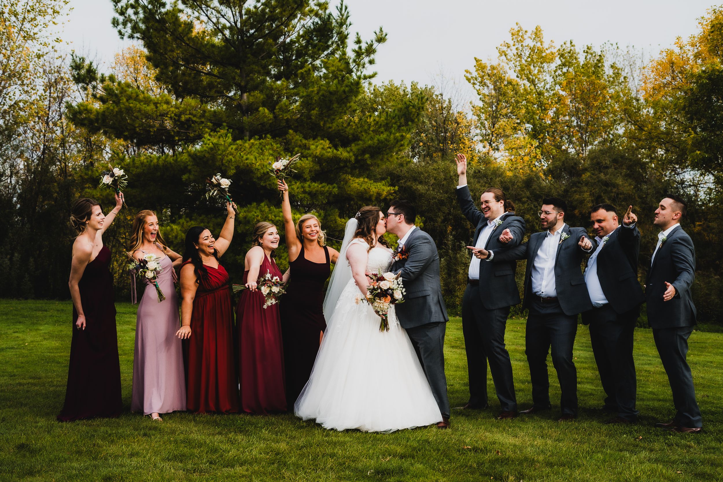 Wedding party poses on lawn with bridesmaids in burgundy dresses and groomsmen in dark suits.