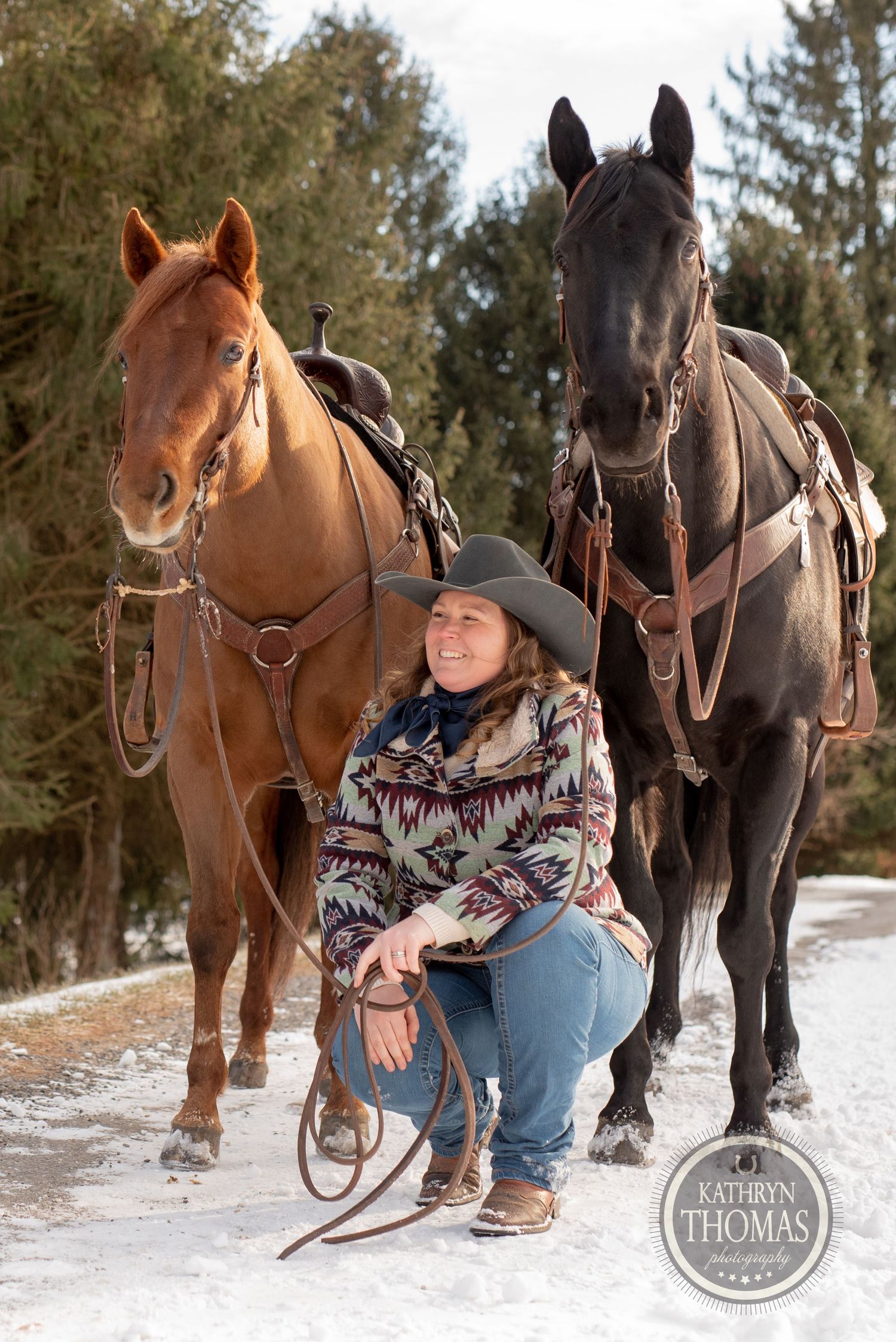 A chestnut and black horse stand saddled in the snow while someone kneels between them.