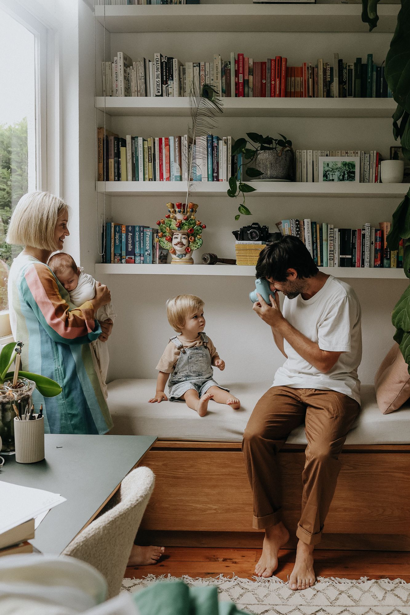 Young family relaxing at home with bookshelves, parents interacting with two toddlers indoors.