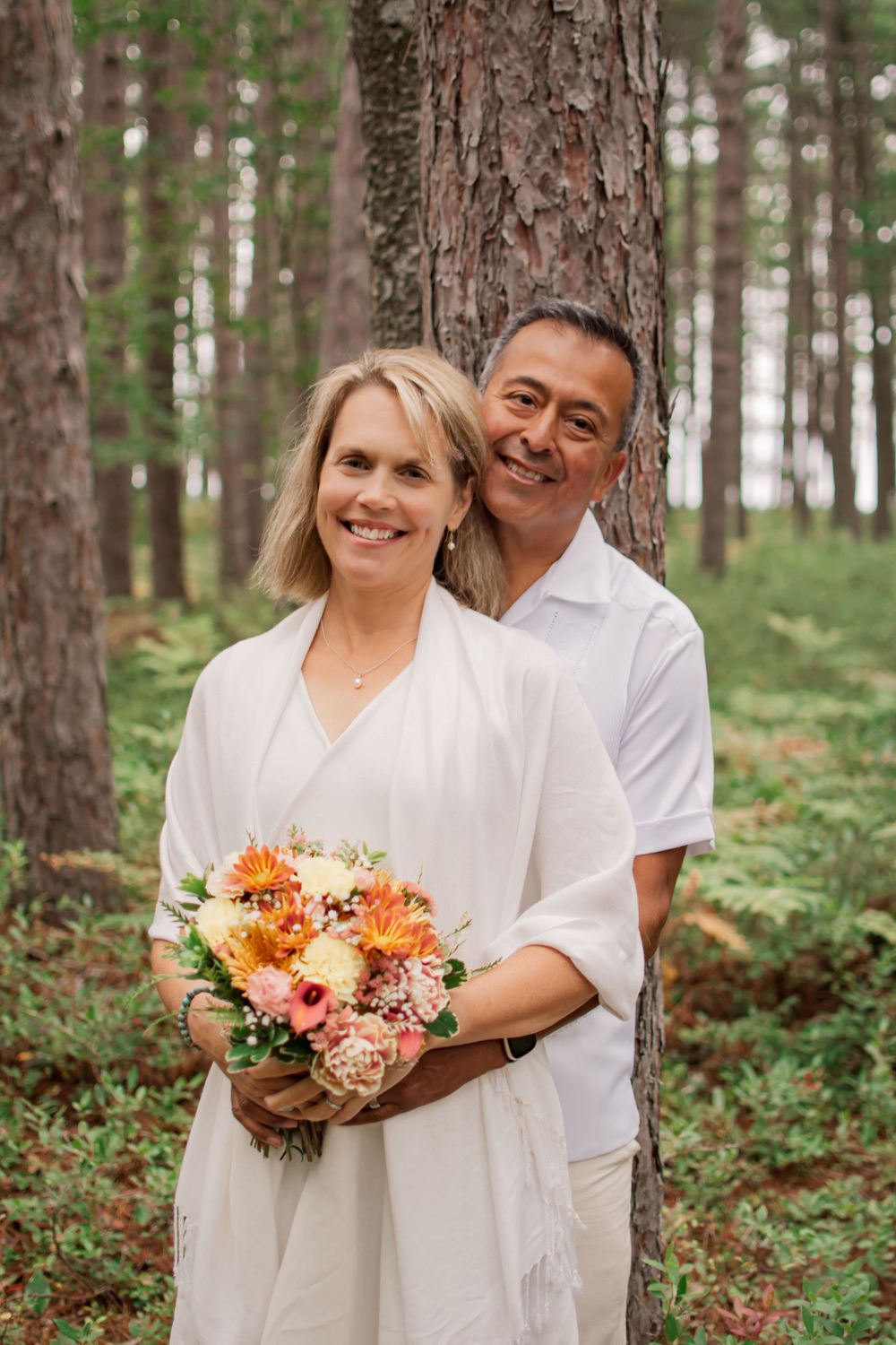 Couple in white clothing poses against pine tree holding colorful floral bouquet.