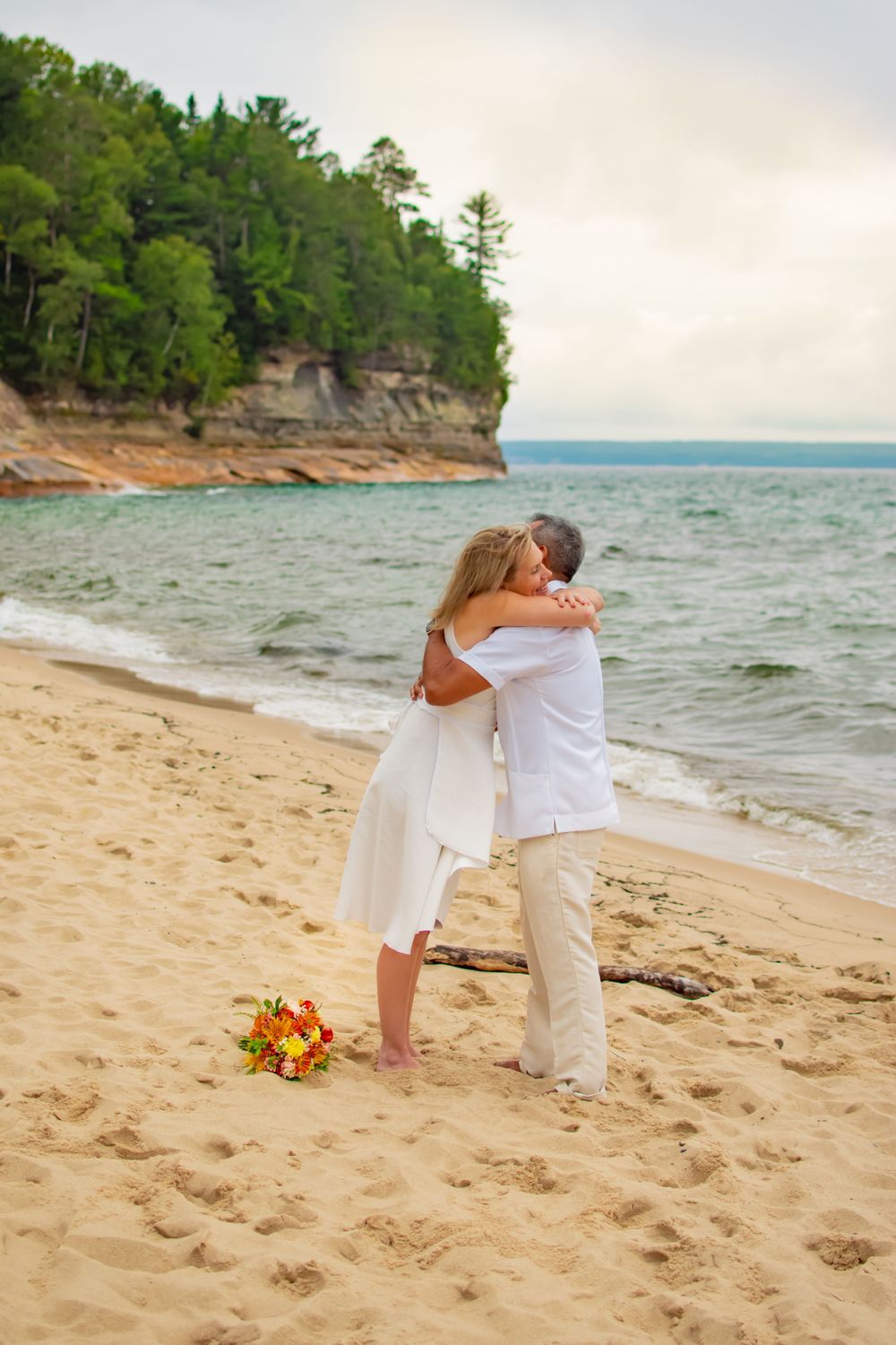 Couple embraces on beach with orange flowers and rocky cliffs in background.