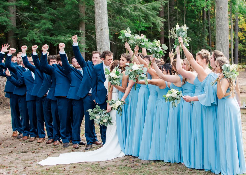 Wedding party in navy suits and light blue dresses pose with raised arms in a forest setting.