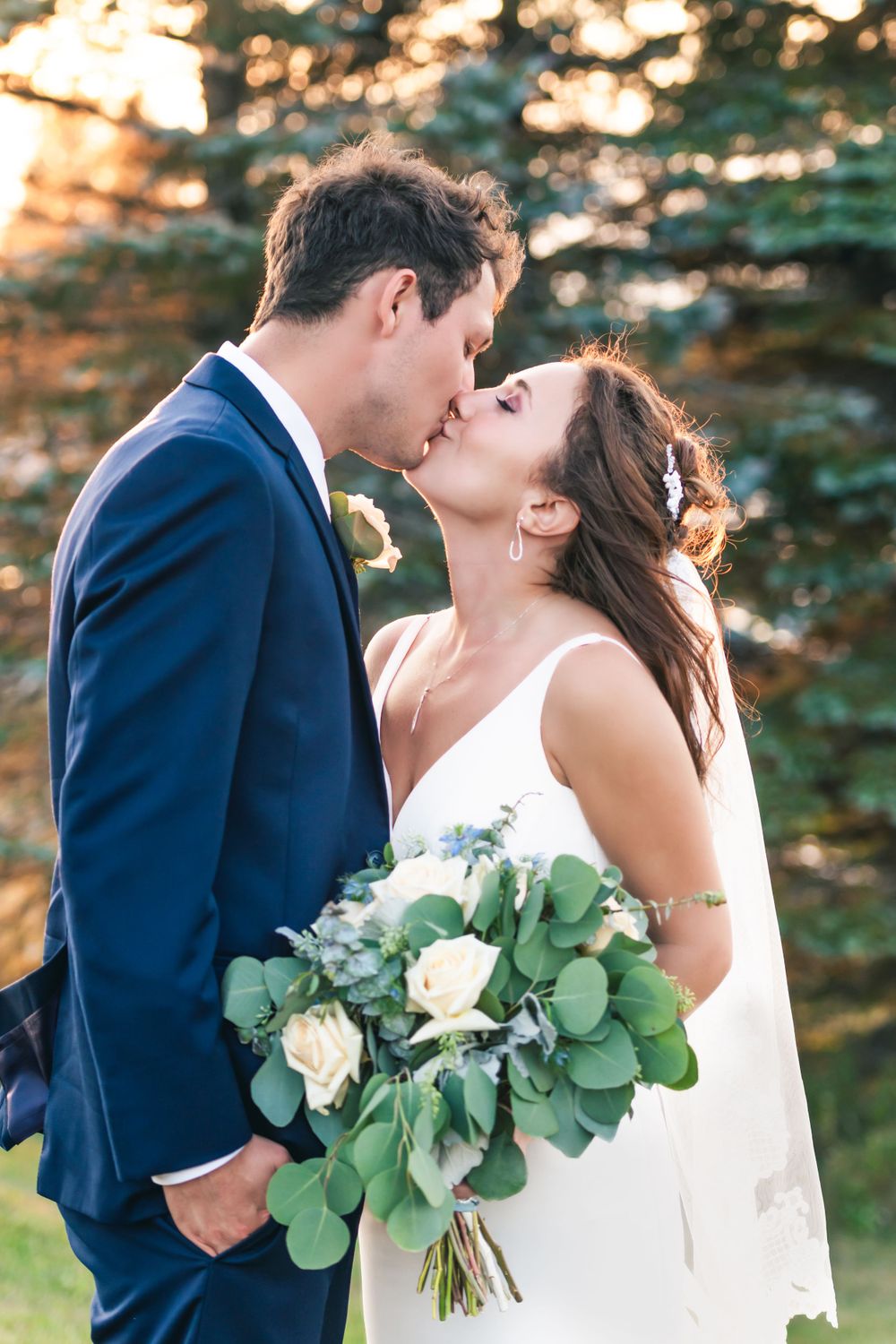 A romantic wedding portrait of a couple kissing with a bouquet of white roses and greenery.