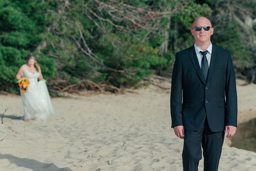 Beach wedding scene with suited groom in foreground and bride visible in background.