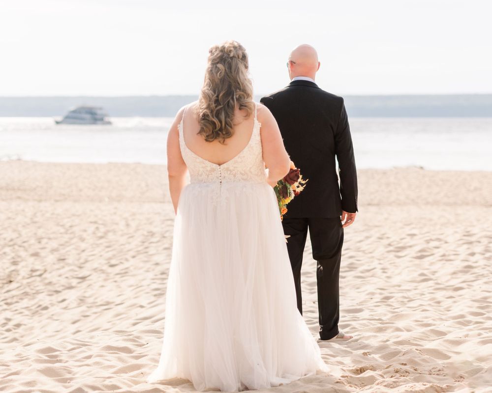 Couple in wedding attire standing on beach facing ocean view.