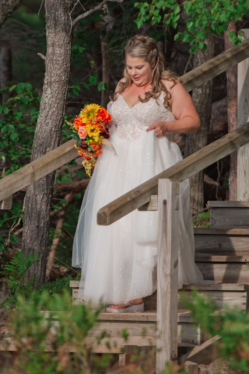 Bride in white gown holding vibrant orange bouquet while descending wooden staircase in forest.