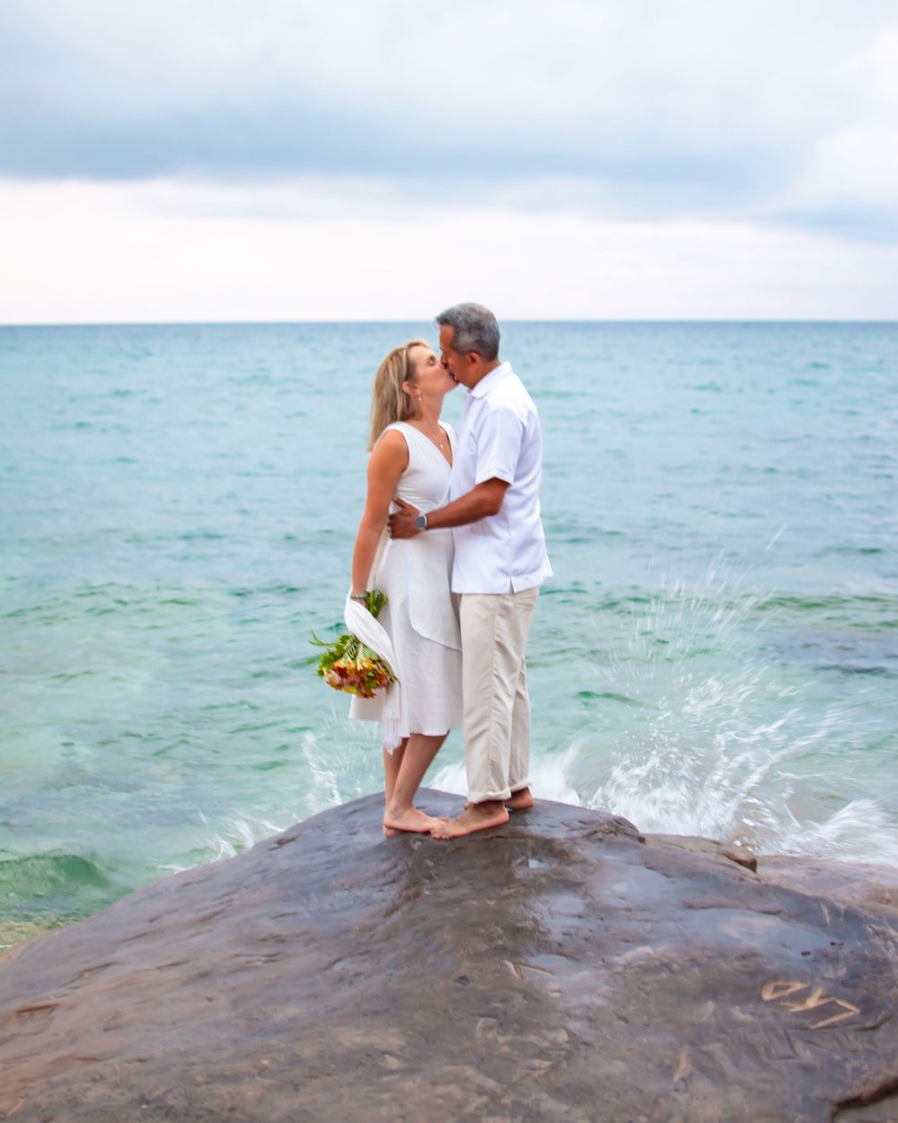 Couple shares romantic moment on rocky outcrop over turquoise waters holding bouquet.