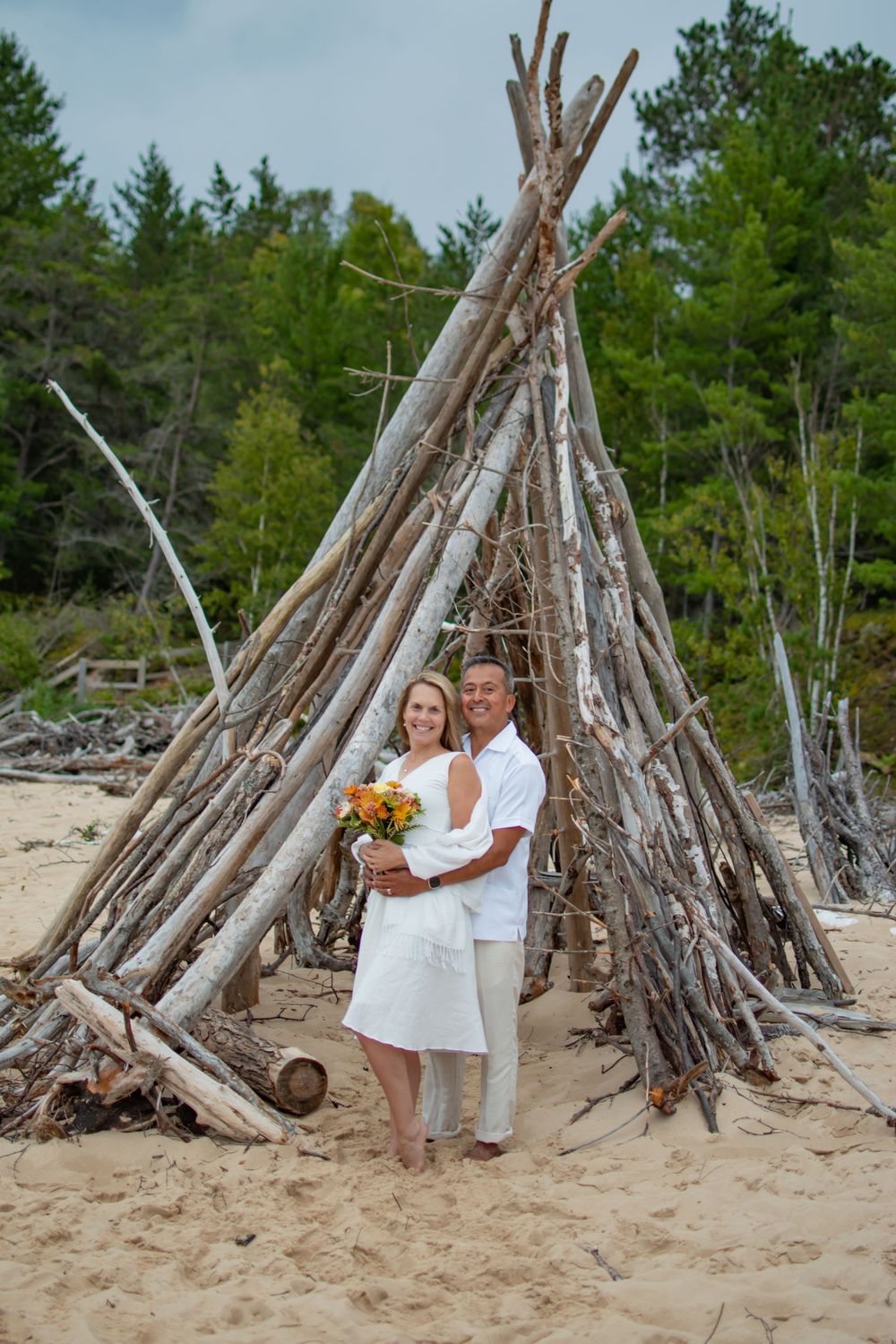 Couple poses inside rustic driftwood teepee structure on beach with pine forest backdrop.