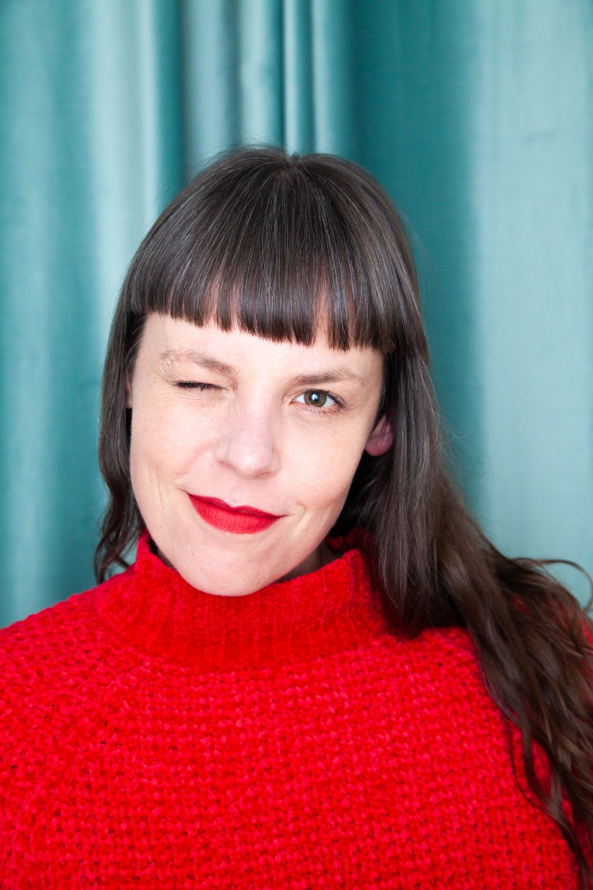 Portrait of person in red turtleneck sweater with bangs against turquoise background.