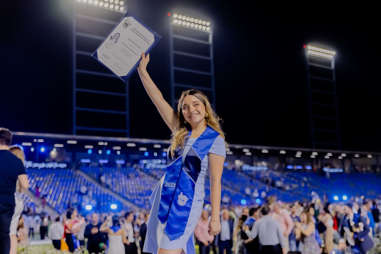 Proud graduate in blue sash holds diploma aloft at night stadium graduation ceremony.