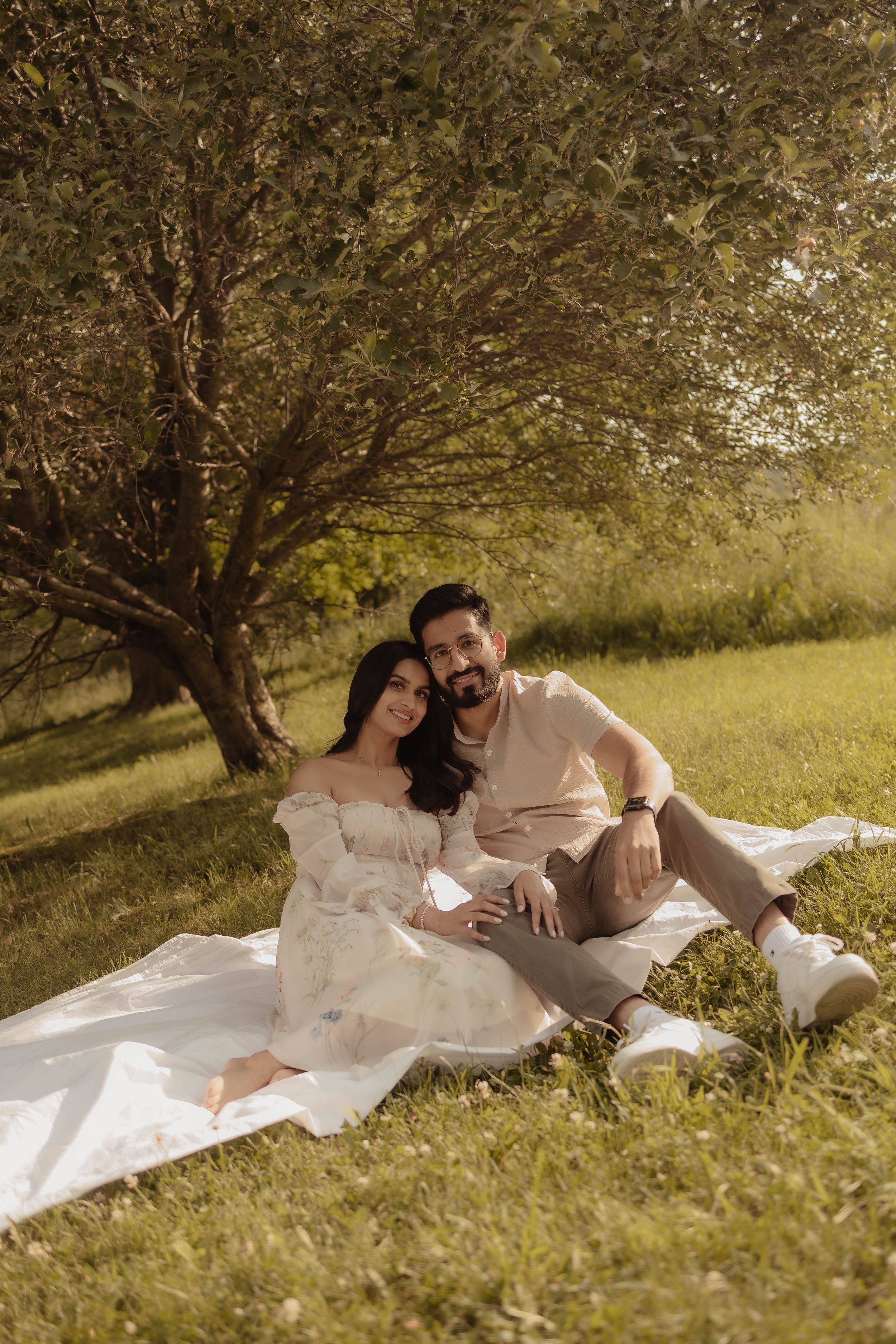 A couple relaxes on a white blanket under a tree in a serene meadow setting.