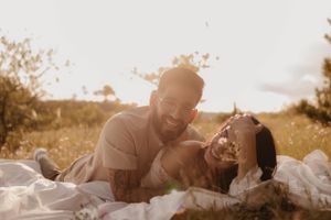 Romantic couple lying together on white blanket in golden sunlit field sharing intimate moment.