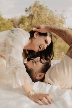 Joyful couple sharing laughter while lying on white blanket in natural setting.