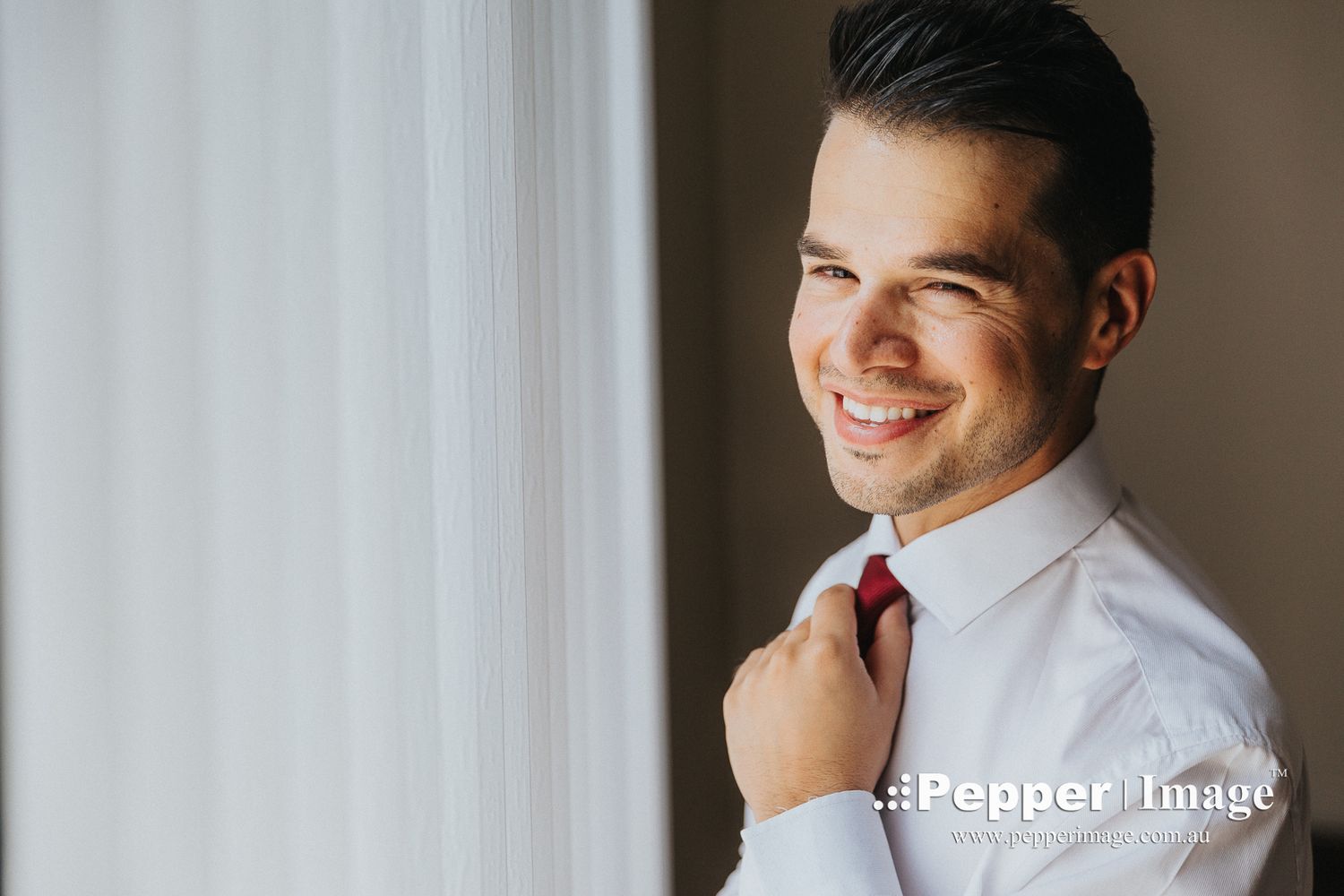 Smiling groom adjusting red tie while getting ready near a bright window on wedding day.
