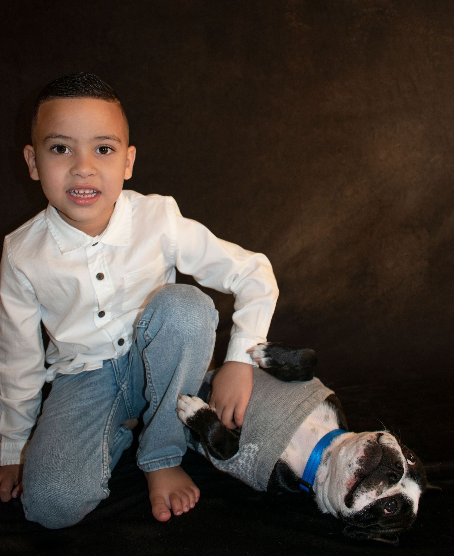 Young boy in white shirt and jeans poses with black and white bulldog on dark studio background.