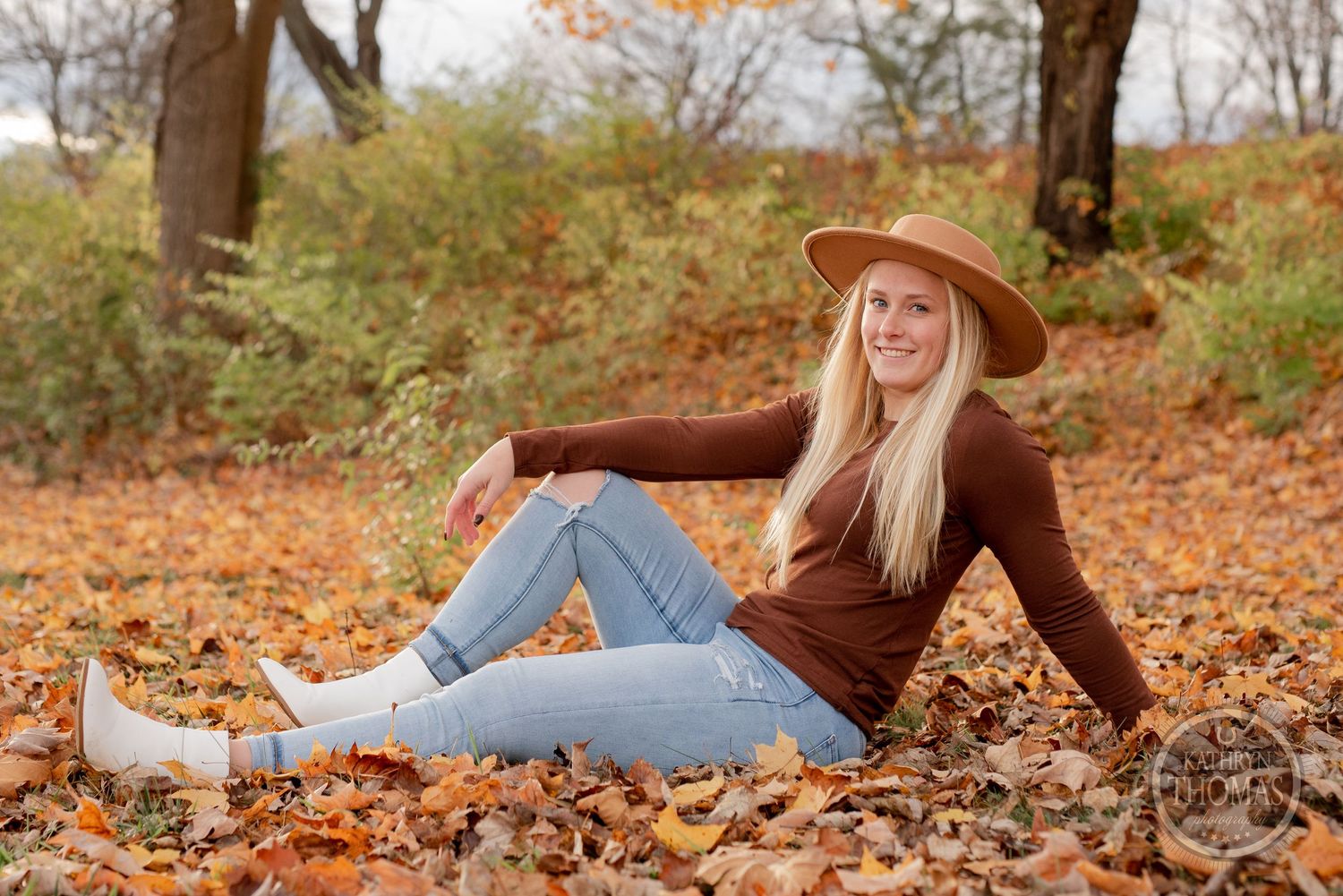 A casual autumn portrait featuring brown sweater and straw hat among scattered fallen leaves.
