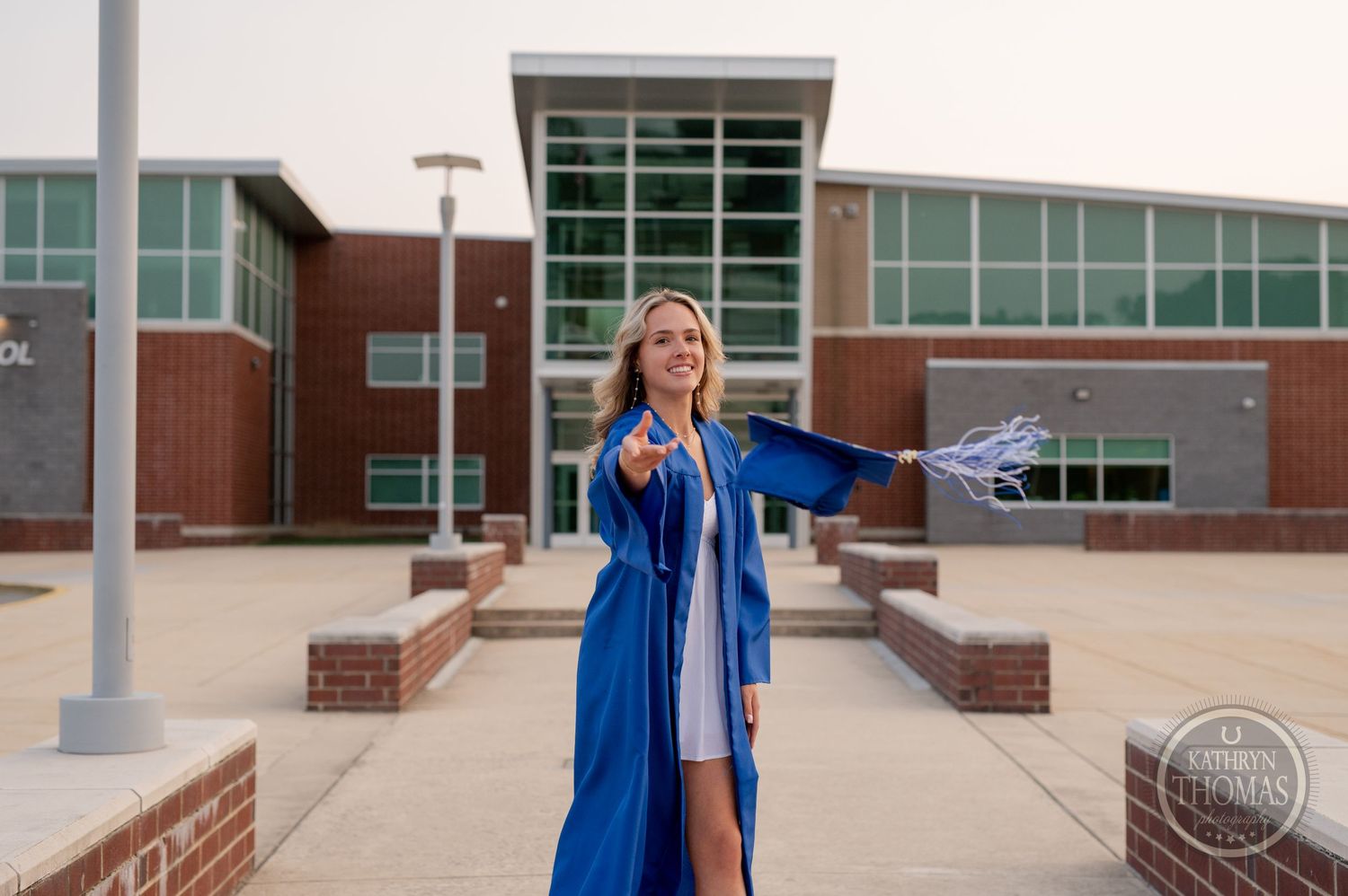 Graduate in blue gown poses with arms outstretched in front of school building.