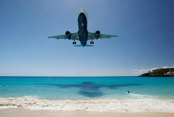 Commercial jet plane flying low over a pristine turquoise beach with crystal clear water and white sand.