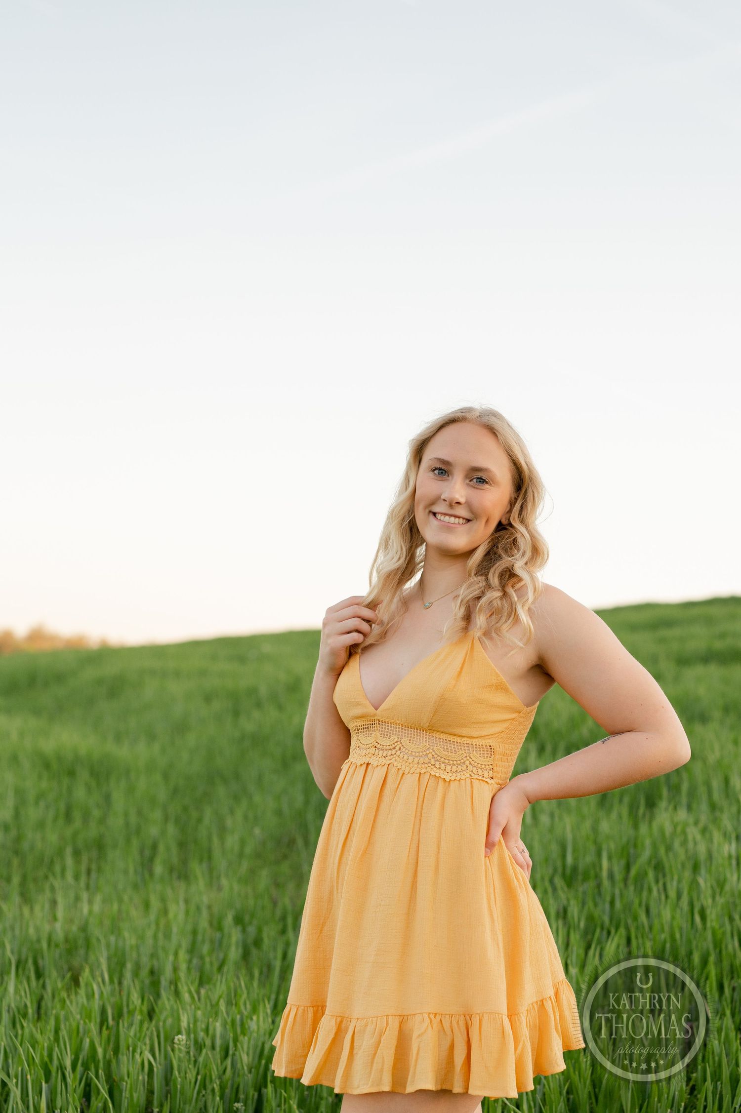 Figure in yellow sundress poses in green spring field during golden hour.