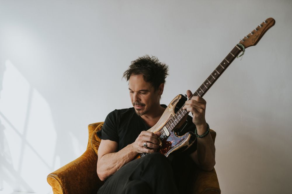 Ian Thornley of Big Wreck sits in a yellow chair with his guitar, he plays a chord looking off to his left.