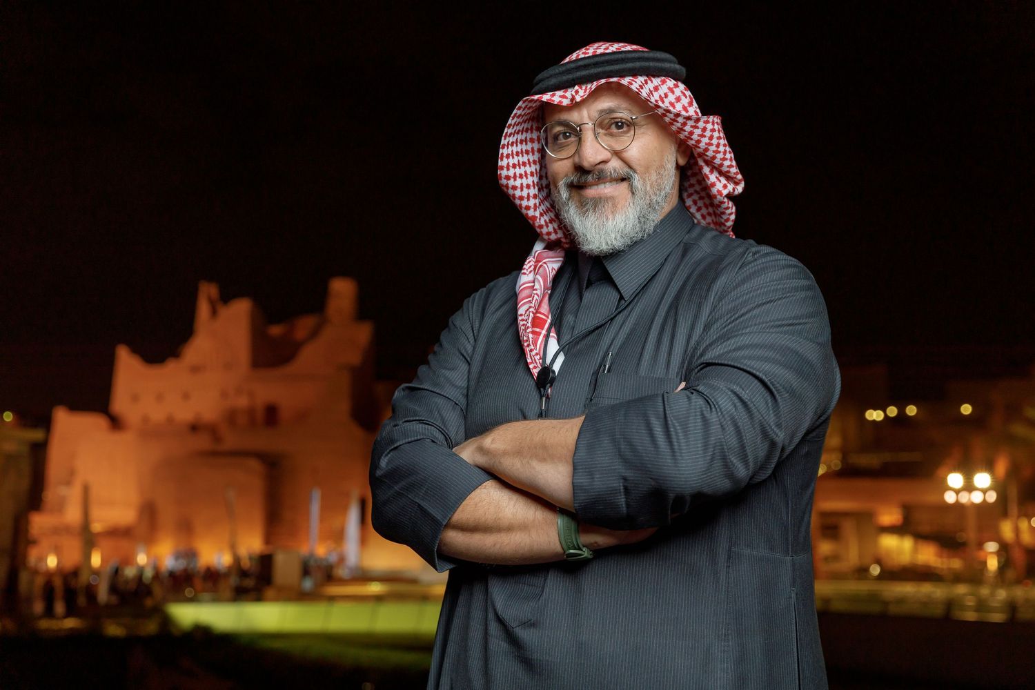 Traditional Middle Eastern dressed man poses confidently at night with illuminated buildings in background.