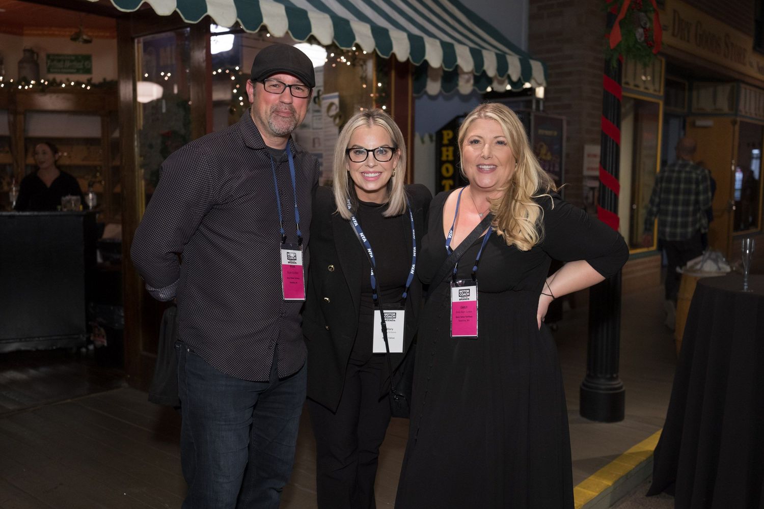 Three people with staff badges pose together backstage at an event venue under a striped awning.