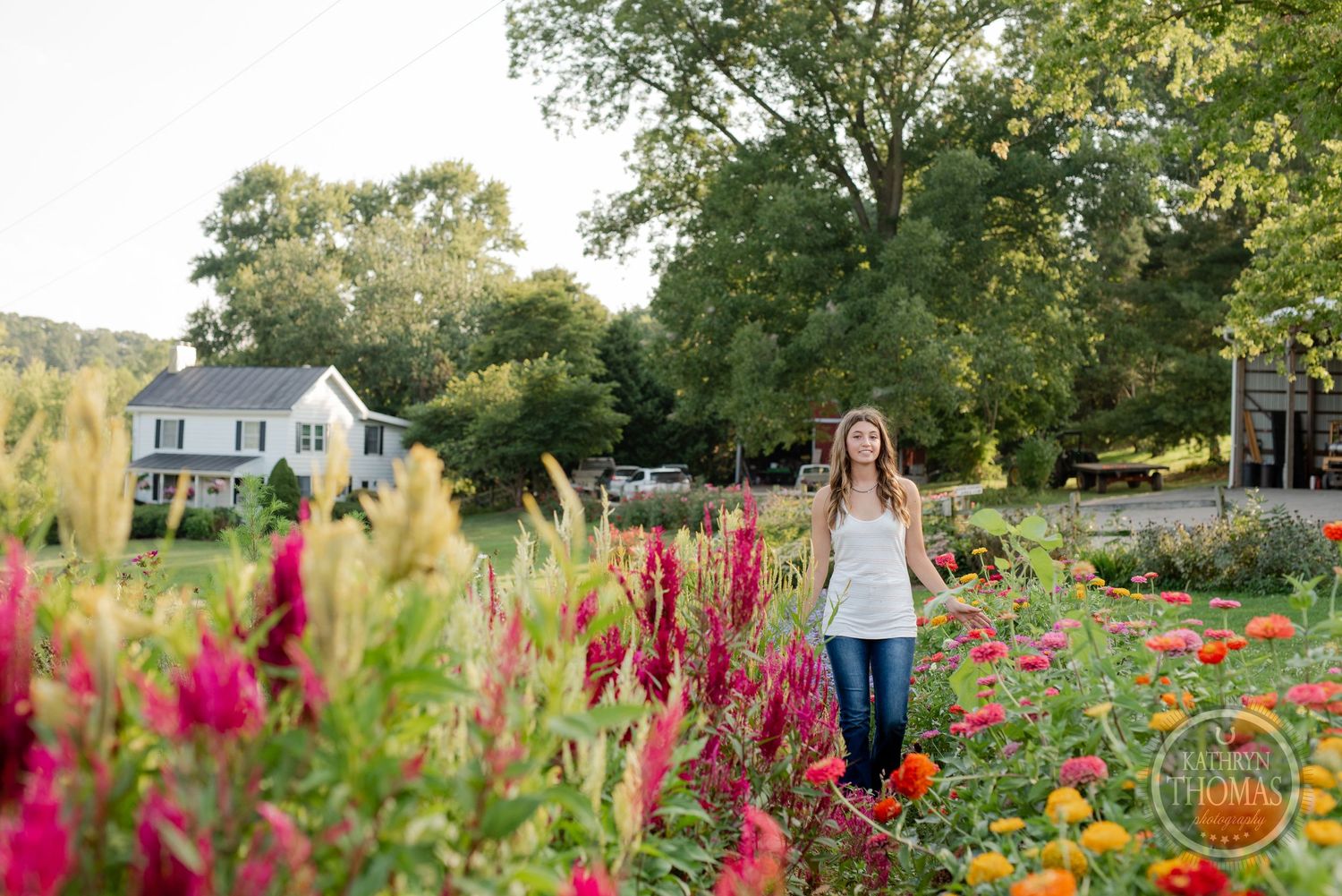 Person standing among tall flowers in a garden with white farmhouse and trees in background.