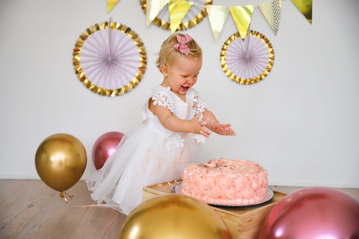 Small child in white dress reaches for pink birthday cake with gold and pink balloons and decorations.
