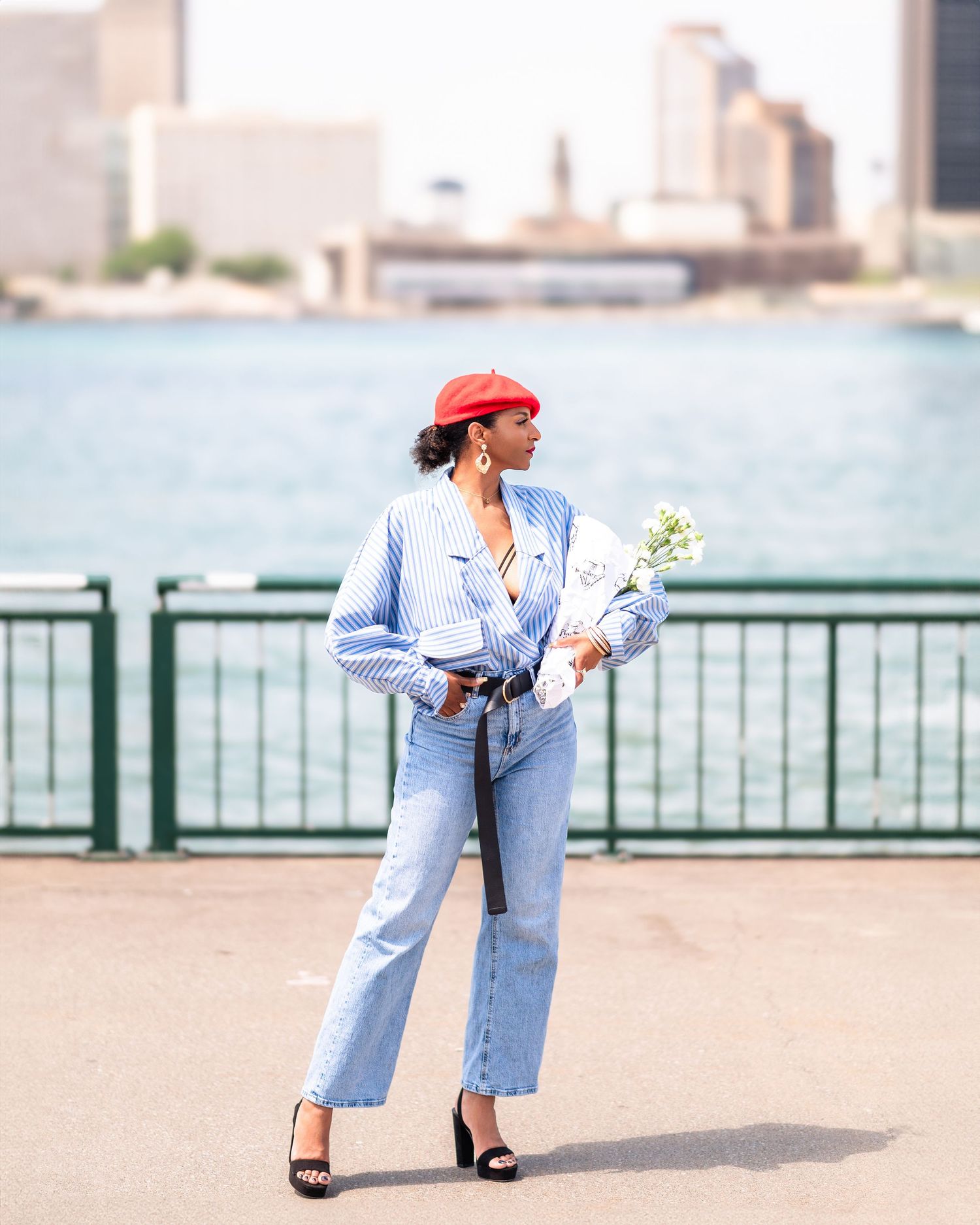 A stylish woman in a red beret and blue outfit poses with flowers along a waterfront.
