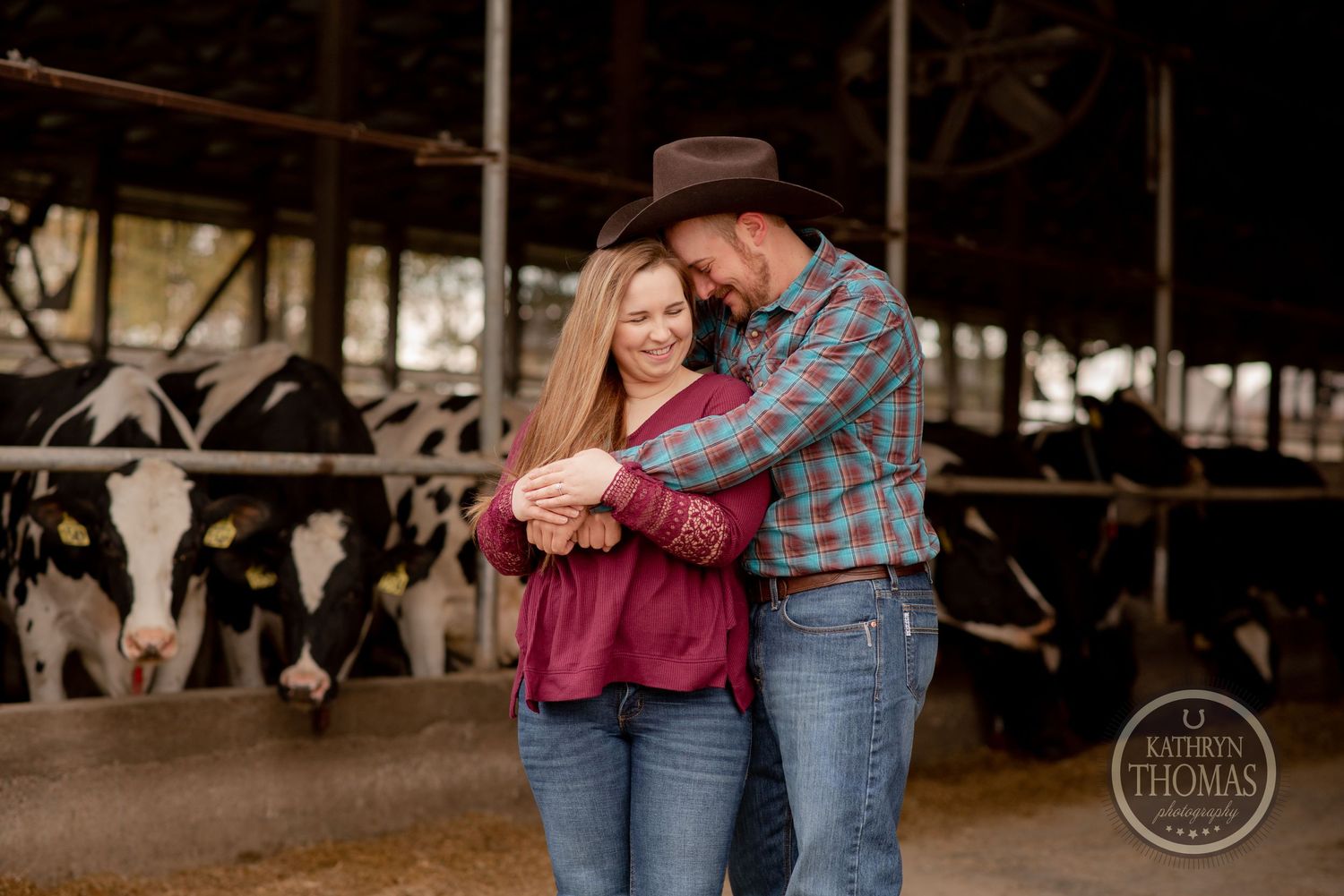 Couple shares intimate moment in barn wearing western attire with cattle stalls behind them.