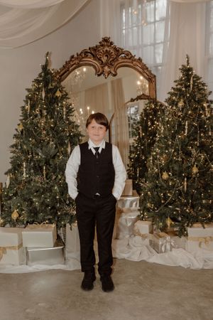 A child standing between two illuminated Christmas trees in formal attire with an ornate mirror backdrop.