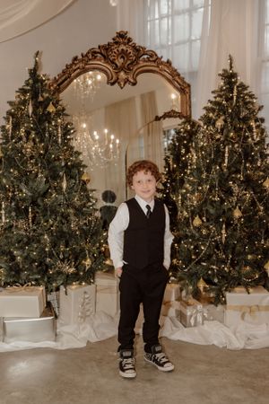 Young boy in formal attire standing between decorated Christmas trees with ornate mirror backdrop.