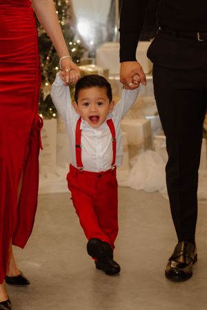 A joyful toddler in red pants and suspenders walking while holding hands during a Christmas photoshoot.