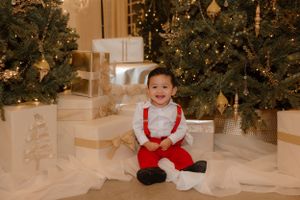 A child in red and white holiday attire sits among decorated Christmas trees and wrapped presents.