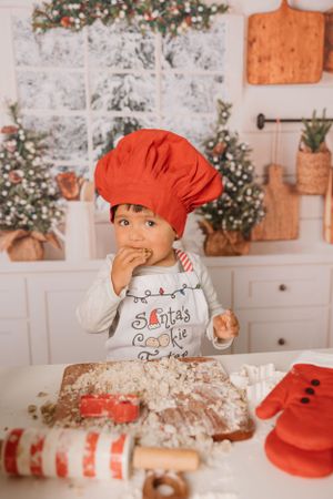 Child in red chef hat and Christmas shirt tasting cookie dough in festive kitchen setting.