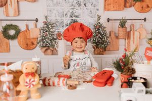 Child wearing red chef hat baking Christmas cookies with holiday decorations in background.