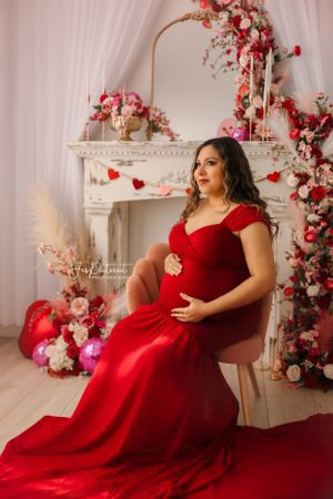 Stunning maternity session with red gown against decorative fireplace with floral arrangements.