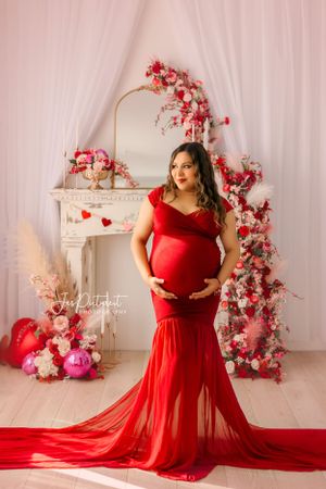Maternity portrait with woman in flowing red dress against pink floral backdrop.