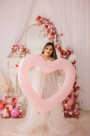 Maternity photo with person holding large pink heart balloon against floral backdrop.