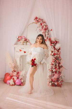Romantic maternity photo featuring white dress and pink floral arch with Valentine's Day decorations.