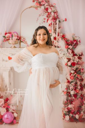 Elegant maternity portrait with pink and red floral backdrop and white gown.